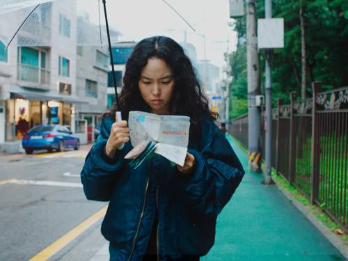 Eine Person läuft die Straße entlang und liest dabei einen Stadtplan. Sie hält außerdem einen durchsichtigen Regenschirm.